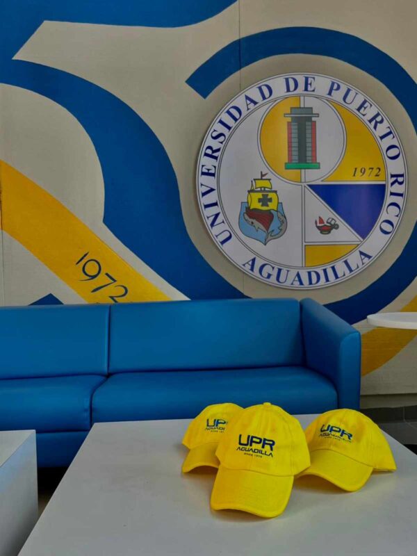 gorras amarillas con logo UPR Aguadilla en azul sobre una mesa con un fondo del logo del 50 aniversario