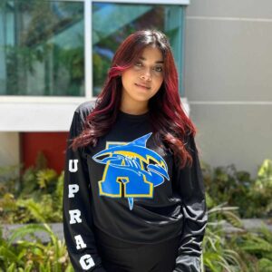 joven dama modelando playera negra con logo de tiburón y letra A en color azul y amarillo en la manga letras UPRAG