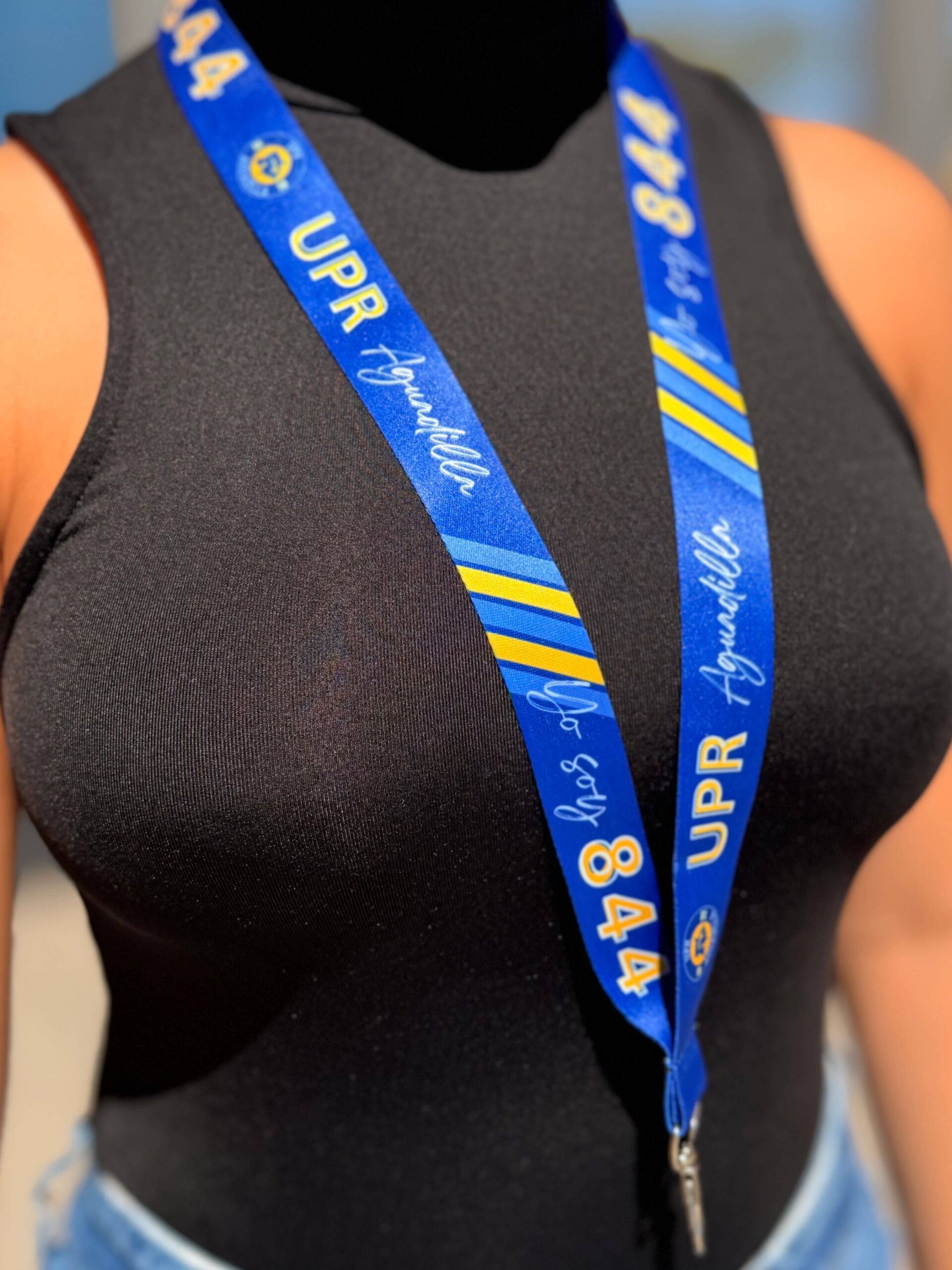 Llavero tipo "landyard" color azul royal con letras amarillas que dicen “UPR Aguadilla” y Yo soy “844”, con un gancho metálico en la parte inferior colgado en el cuello de una persona.