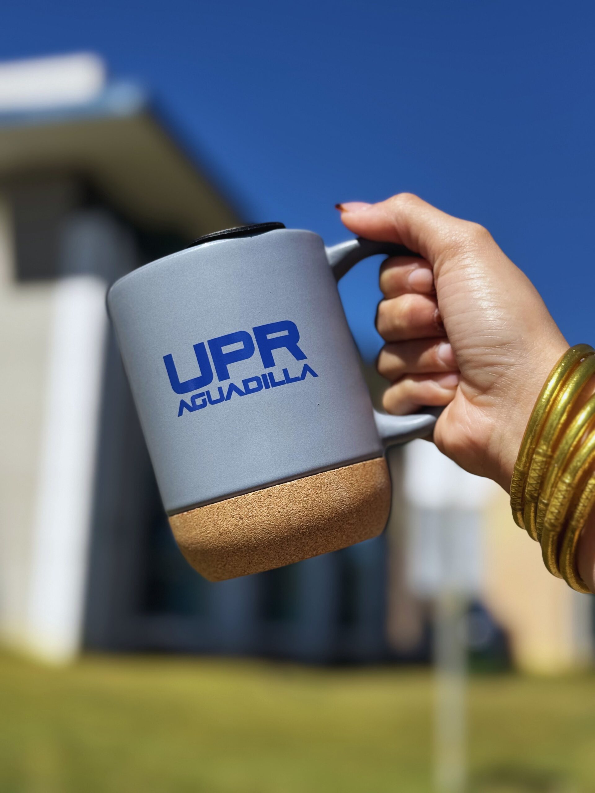 Taza gris con el logo UPR Aguadilla en color azul royal; la taza tiene base de corcho y tapa negra.