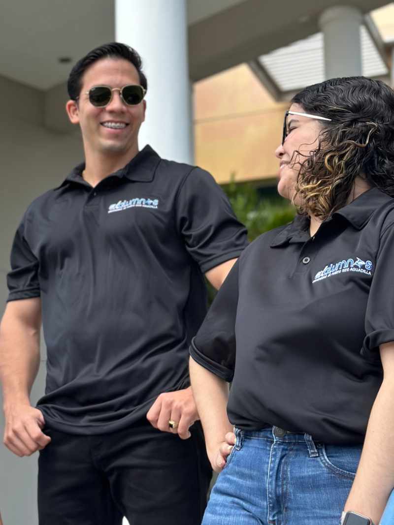 Polo en microfibra color negra con bordado de exalumnos UPR Aguadilla en color azul con detalles blancos y amarillos en el borde, colocado en el lado izquierdo, modelado por una mujer y un hombre en el exterior de la biblioteca.