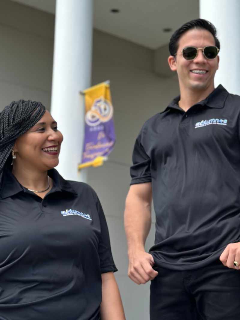 Polo en microfibra color negra con bordado de exalumnos UPR Aguadilla en color azul con detalles blancos y amarillos en el borde, colocado en el lado izquierdo, modelado por una mujer y un hombre en el exterior de la biblioteca.