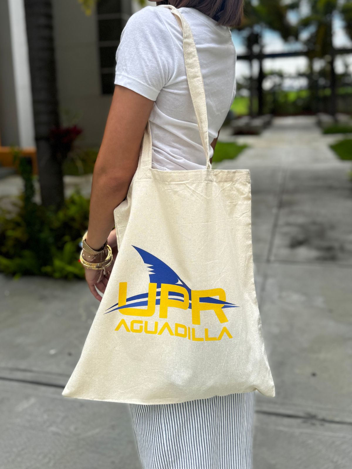Bolso tipo tote color beige con logo estampado de UPR Aguadilla en color amarillo con aleta de tiburón color azul royal entrelazada en las letras, lo lleva una joven en el hombro.