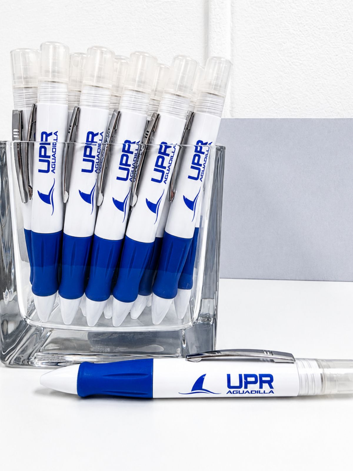 Bolígrafos promocionales UPR Aguadilla en colores blanco y azul, exhibidos en un recipiente de vidrio con un bolígrafo en primer plano.