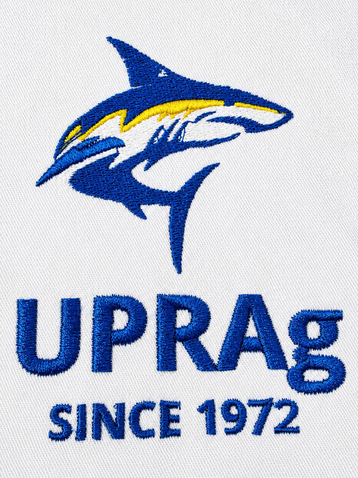 Logo bordado de UPR Aguadilla con tiburón azul y detalles en amarillo sobre tela blanca, acompañado del texto "Since 1972".