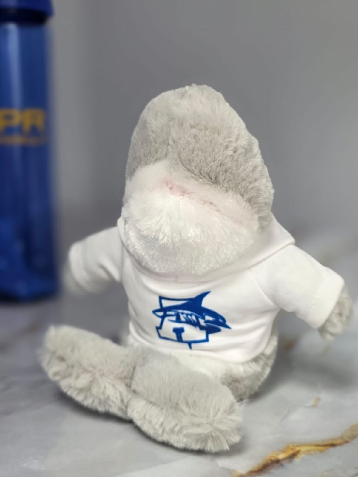 Peluche de tiburón gris con sudadera blanca estampada con el logo de UPR Aguadilla en azul royal, sobre superficie de mármol con botella institucional desenfocada al fondo. Enfoque en área frontal para distinguir logo