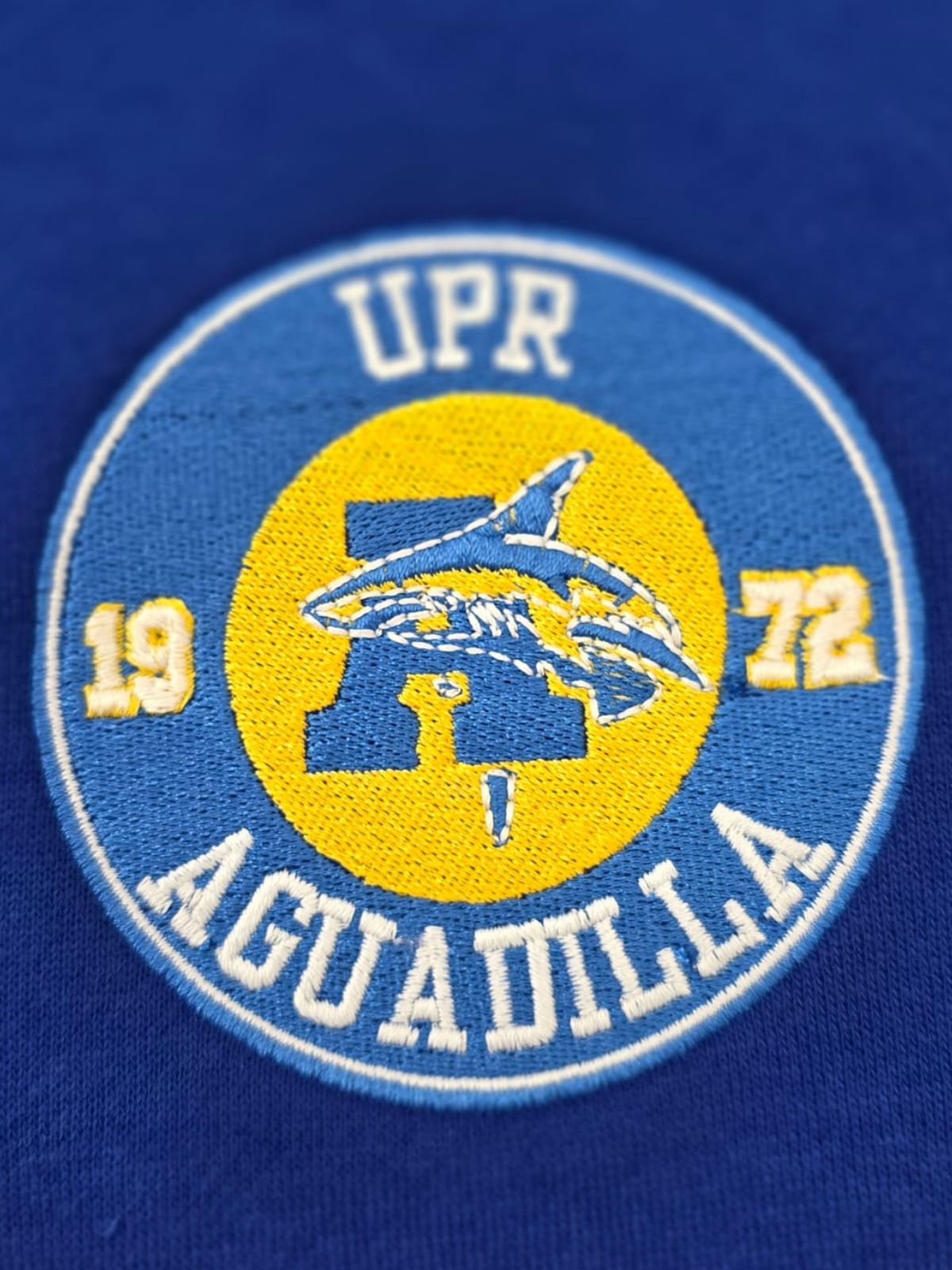 Sello totalmente bordado redondo en color azul con centro amarillo y letra A con tiburón en azul. En el borde redondo UPR Aguadilla 1972 en blanco.