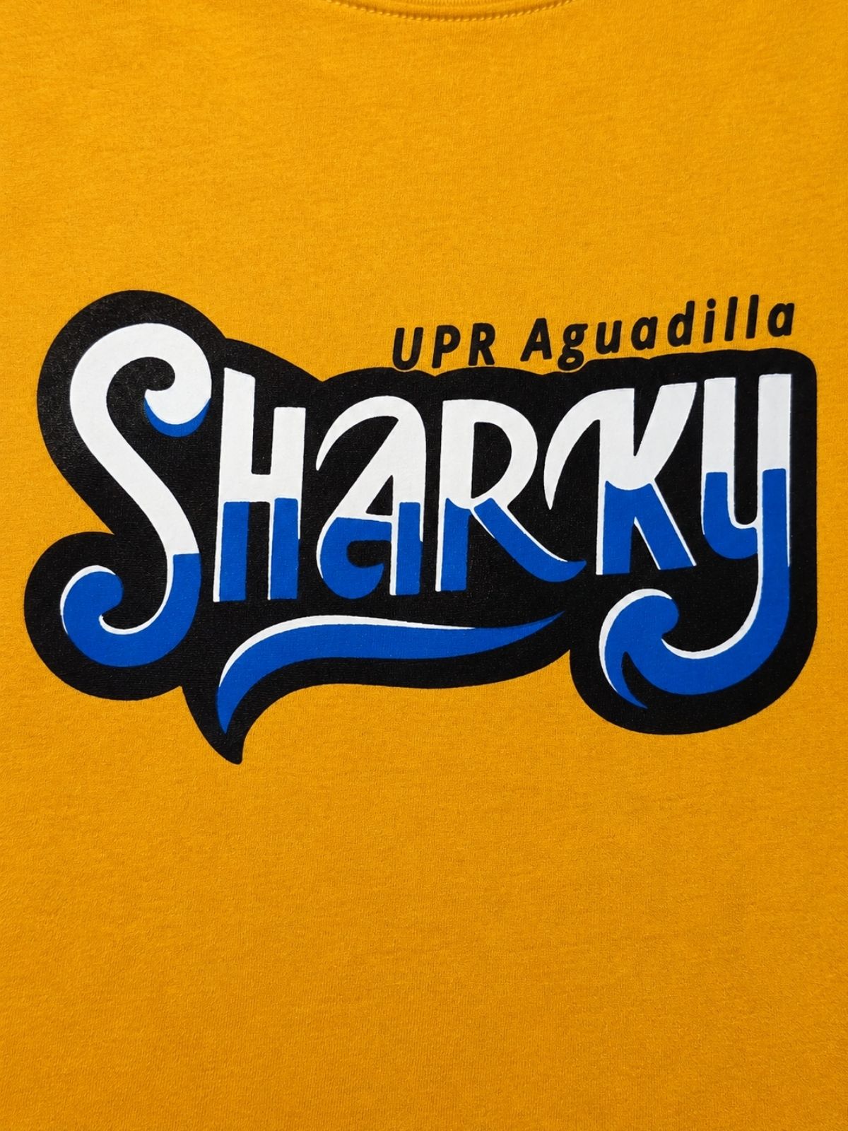 Detalle de estampado de camisa amarilla Sharky UPR Aguadilla,, estampado en color amarillo y blanco con fondo negro sobre tela amarillo