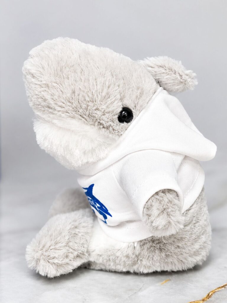 Peluche de tiburón representativo de la mascota institucional en color gris claro, tiene el pecho blanco, ojos en forma de botones color negro. Está vestido con un abrigo blanco y estampado de la letra insignia con tiburón en color azul royal.